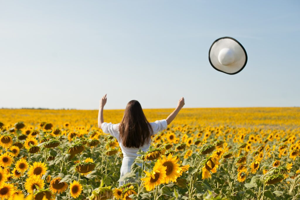 Woman in sunflower field on a sunny day with blue sky, young, happy, free, hands and hat in the air